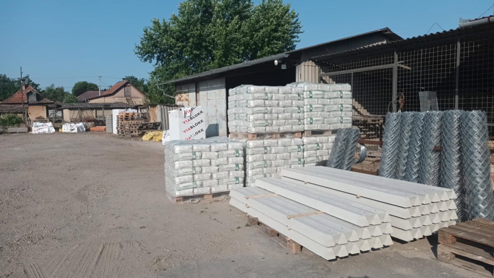 A telep udvara Albertirsán: raklapos cementzsákok, betonoszlopok és drótfonat-tekercsek a szín előtt.
